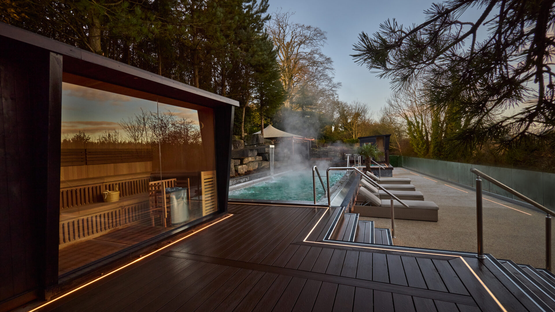 Outdoor cedar sauna with glowing windows beside a steaming plunge pool at dusk — Nore Haus install ambiance